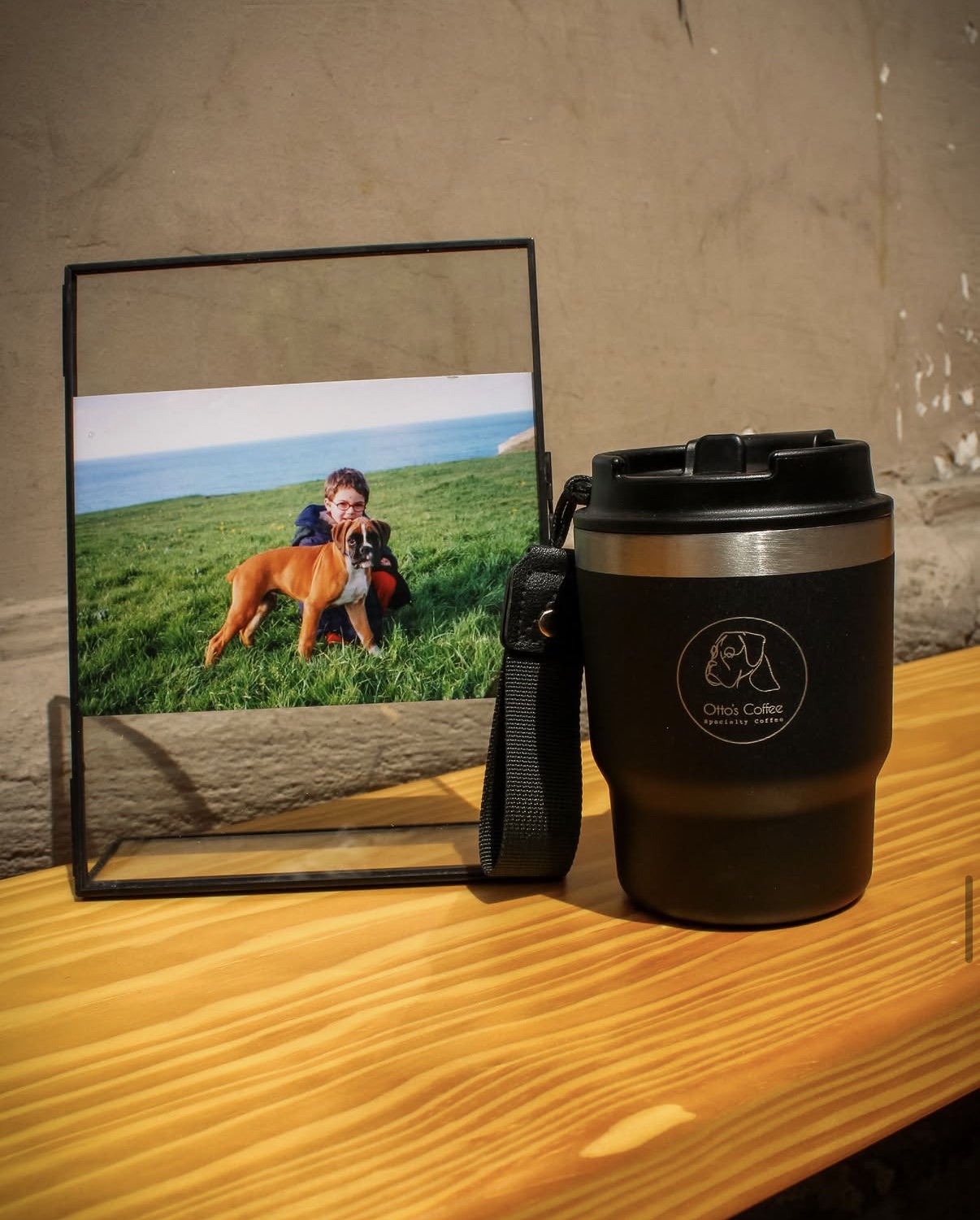 Foto enmarcada de Otto, un bóxer, junto a un niño en la hierba; al lado, un vaso térmico negro con el logo de Otto's Coffee